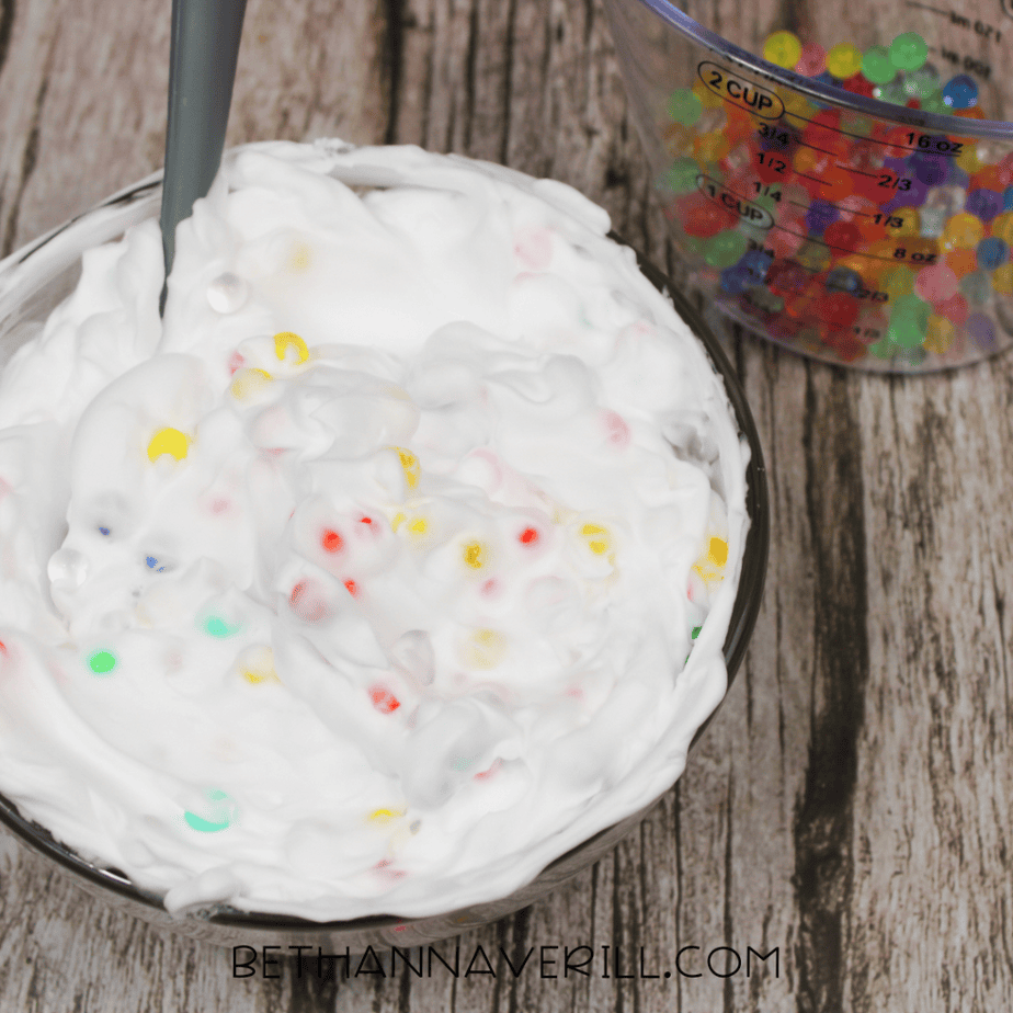 White foam partially mixed with multicolored water beads in a glass bowl, with a measuring cup of additional beads behind it.