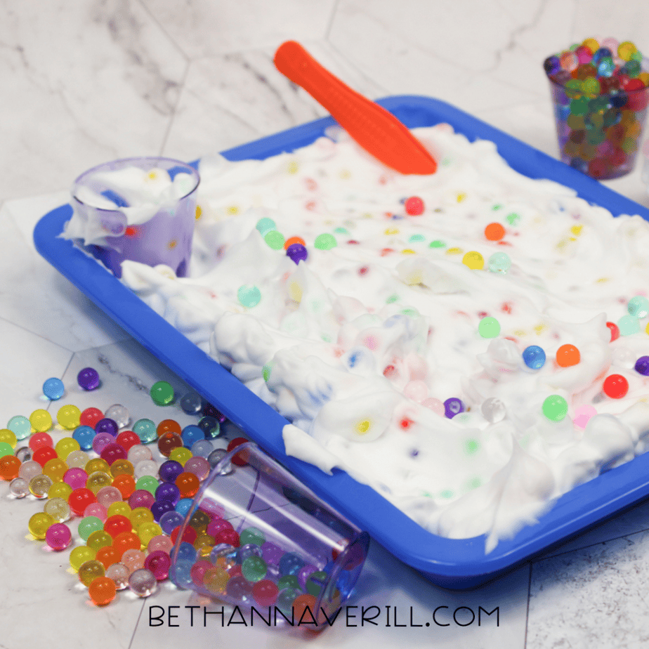 A blue sensory tray filled with white foam and scattered water beads. Small plastic cups and a scoop sit in and around the tray, with extra beads spilled nearby.