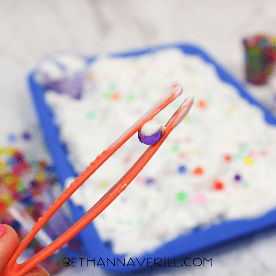 Orange plastic tweezers holding a single purple water bead covered in a bit of white foam, with the sensory tray blurred in the background.