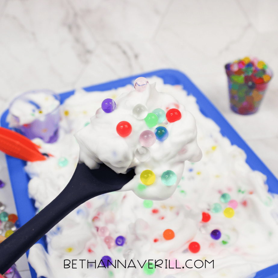 A black spoon holding a scoop of white foam topped with colorful water beads above a blue sensory tray.