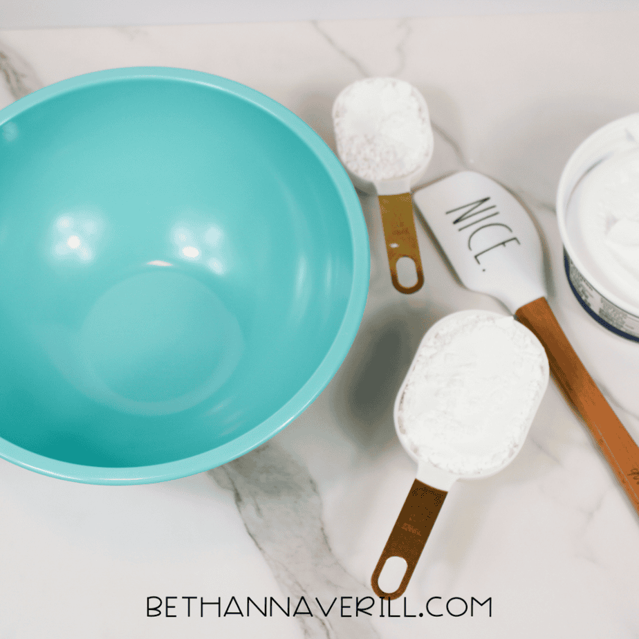 Teal mixing bowl with two white measuring cups filled with white powder and a white spatula on a marble surface.