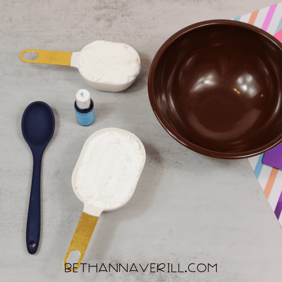 Measuring cups filled with white ingredients, a brown mixing bowl, a spoon, and a small bottle of blue liquid on a countertop.