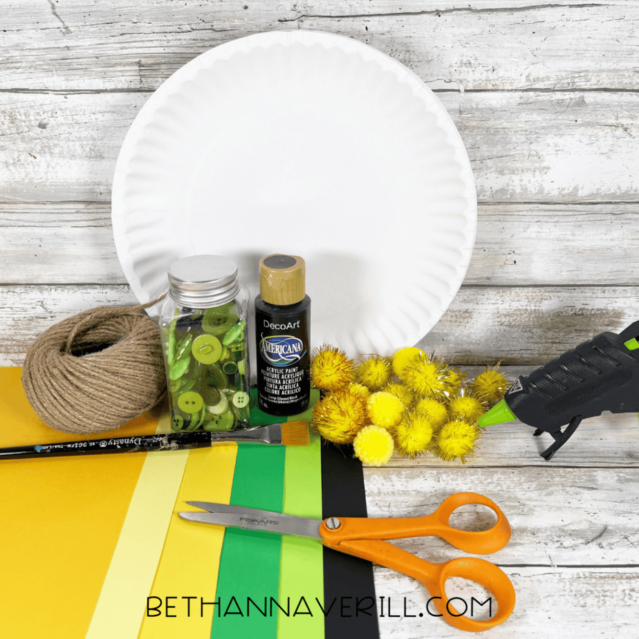 Image of the supplies needed to make a Paper Plate Pot of Gold craft including a white paper plate, black acrylic paint, paintbrush, scissors, glue gun, yellow pom poms, green and yellow buttons, green, black, and yellow cardstock, pencil, and twine arranged on a wood background.