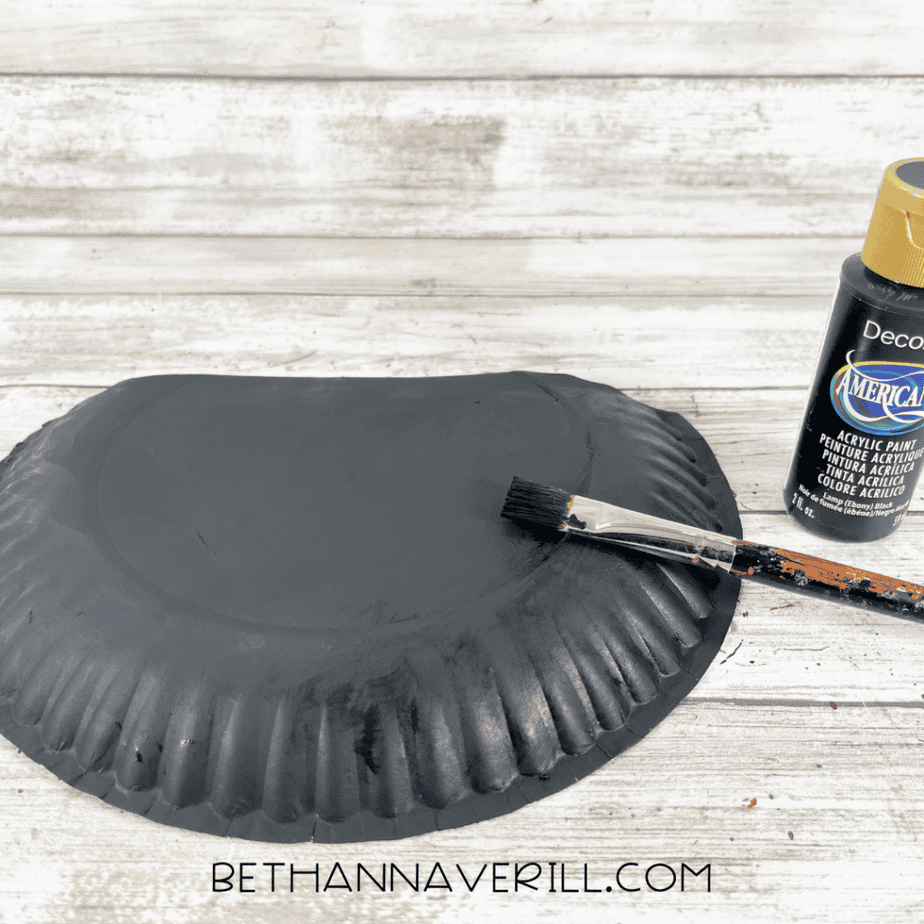 Image of the paper plate painted black with a paintbrush and bottle of black acrylic paint next to it.