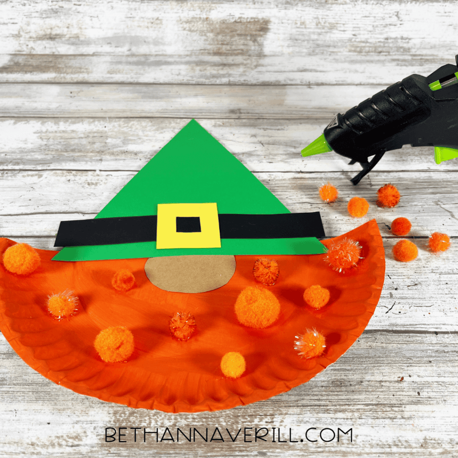 Orange pom poms being placed on an orange painted paper plate beard below a green hat.