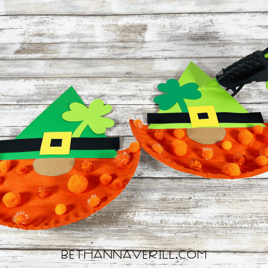Decorative elements being glued to paper plate gnome crafts with green hats and orange pom pom beards on a wood background.