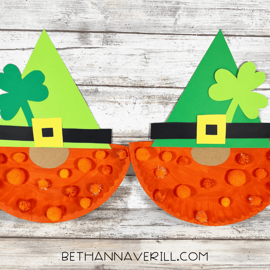 Close up of two completed paper plate gnome crafts with green hats, shamrocks, orange pom pom beards, and yellow buckles.