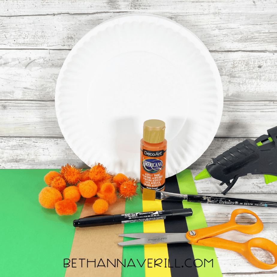 Paper plate, orange pom poms, orange acrylic paint bottle, paintbrush, scissors, marker, glue gun, and sheets of green, yellow, black, and brown cardstock on a wood background.