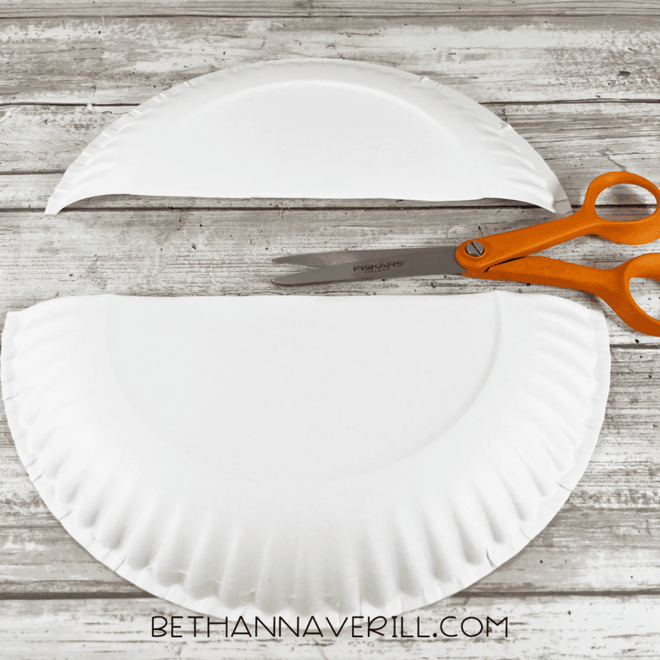 Paper plate cut into two curved pieces with orange scissors beside them on a wood background.