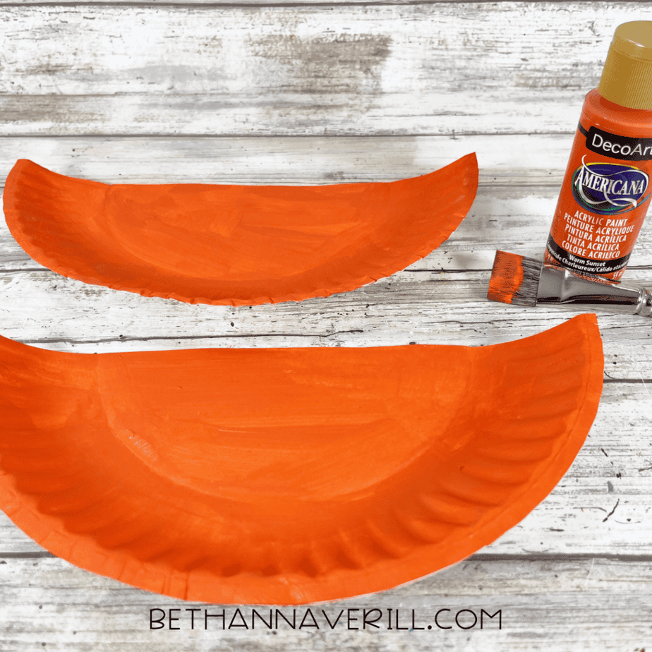 Two curved paper plate pieces painted orange with an acrylic paint bottle and paintbrush nearby.