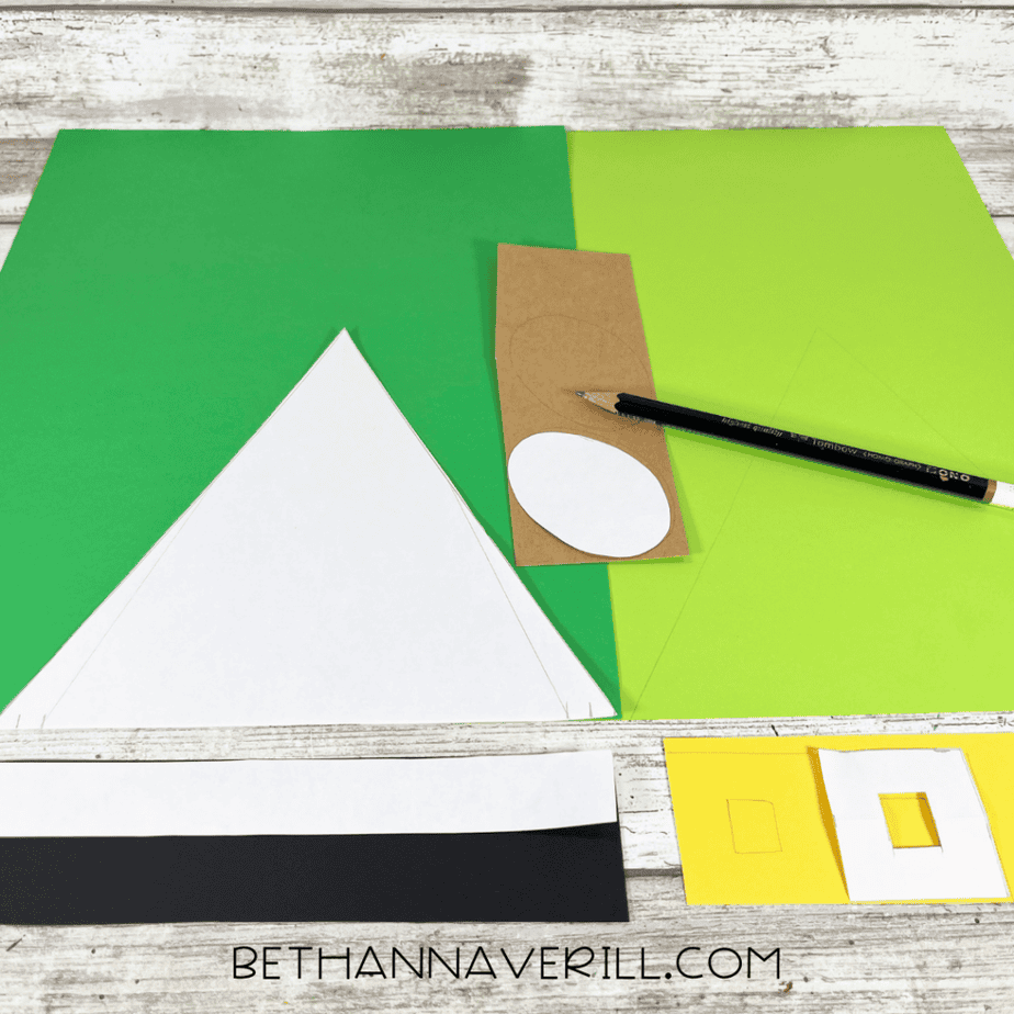 Green cardstock triangles, shamrock cutouts, black hat bands, yellow buckle shapes, and brown circle pieces arranged on a wood surface with scissors.
