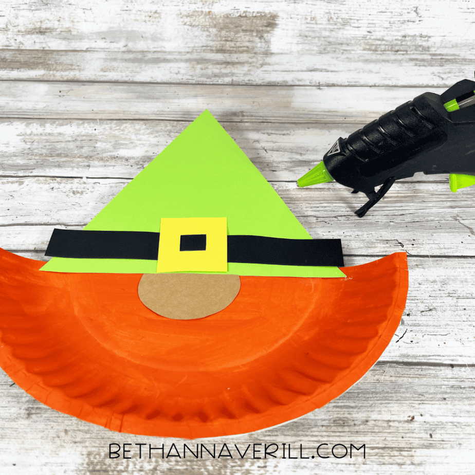 Paper plate piece with light green cardstock hat attached and a glue gun positioned nearby.