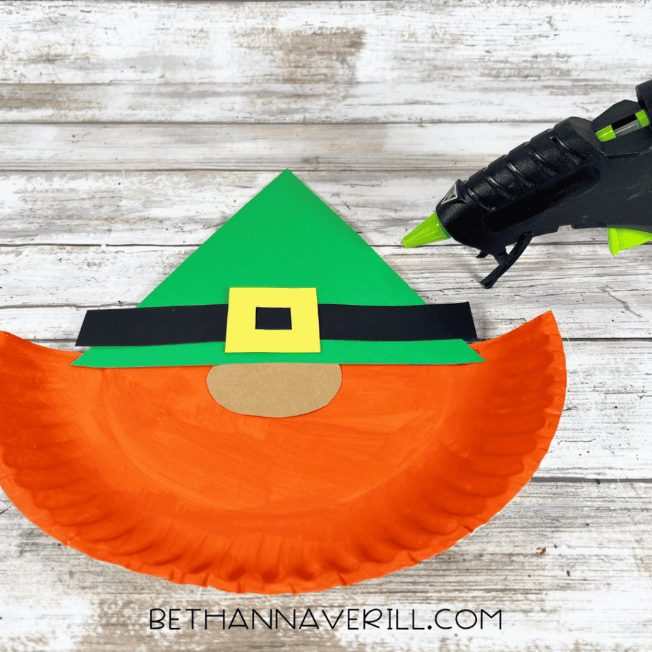 Paper plate piece with dark green cardstock hat attached and a glue gun positioned nearby.