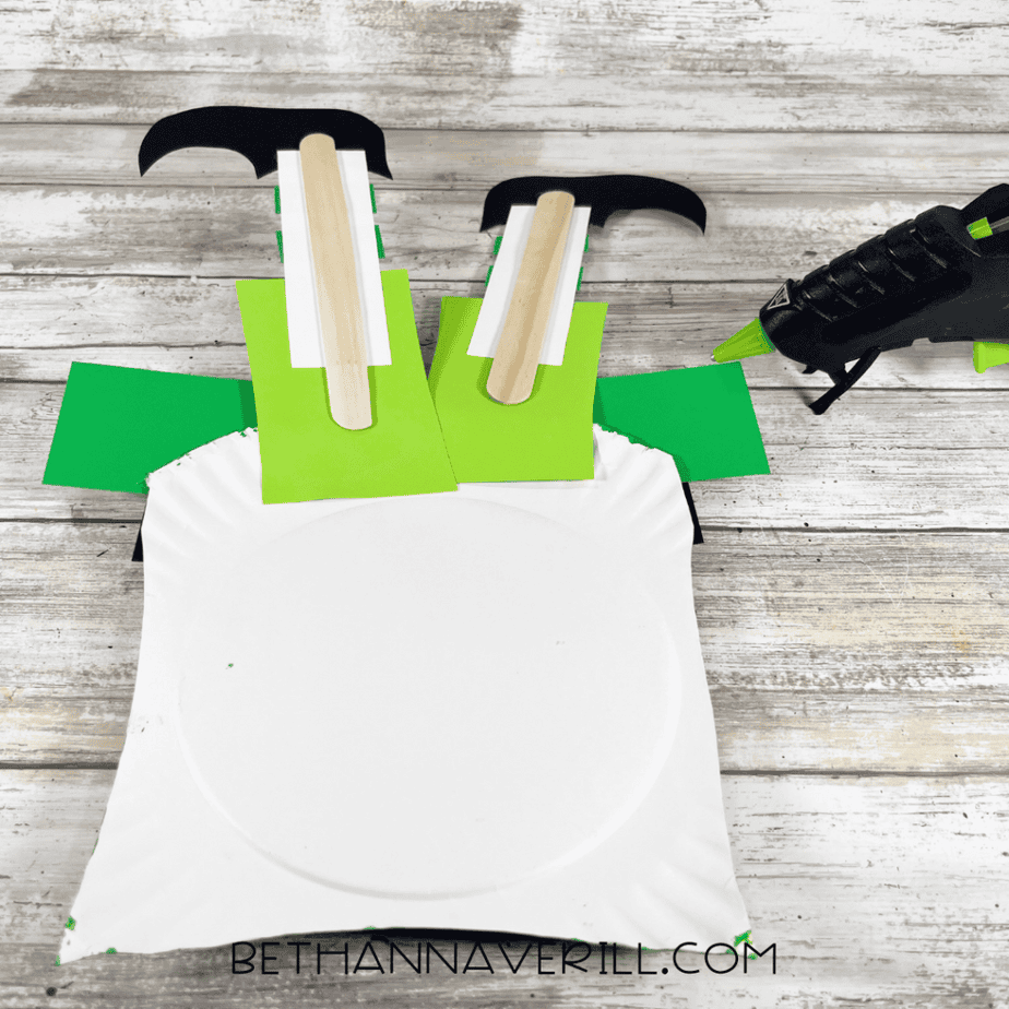 A painted green paper plate piece with a black cardstock strip and yellow square frame attached to the front and a shamrock shape on the side.