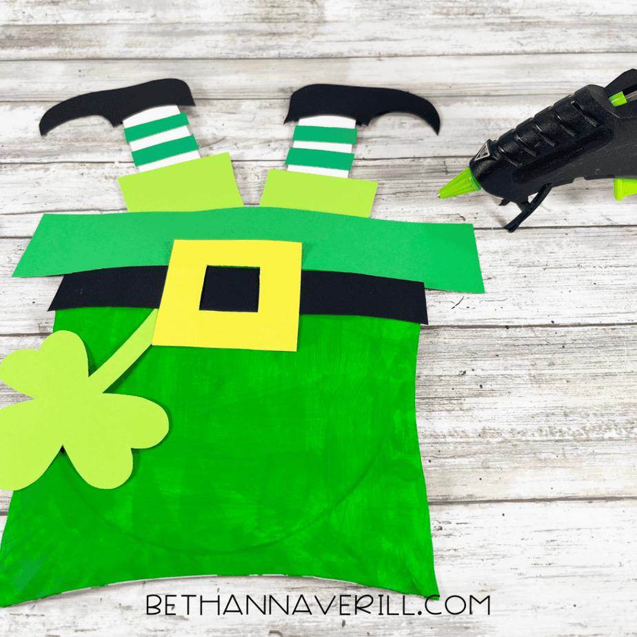 Paper plate craft showing two striped legs with black shoes sticking up from a green paper plate piece with a shamrock attached.