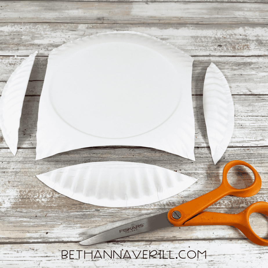 A white paper plate cut into three curved pieces placed on a wooden surface next to orange scissors.