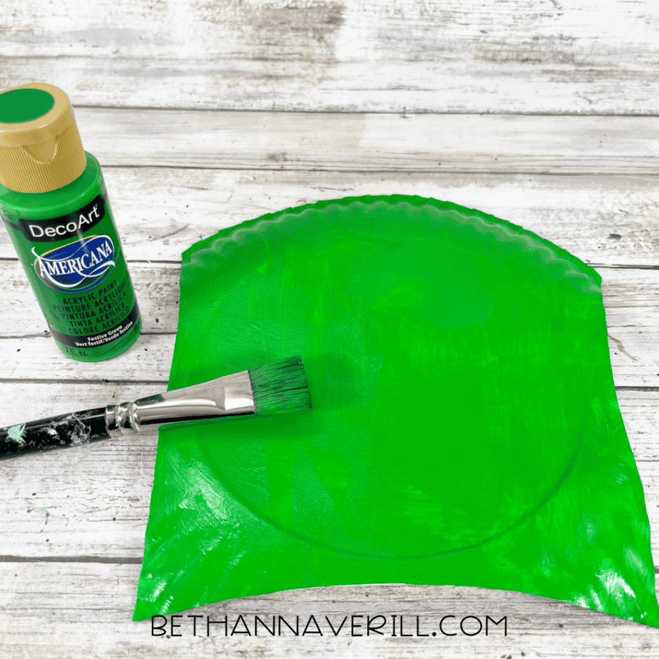 A cut section of a paper plate being painted bright green with acrylic paint using a paintbrush.