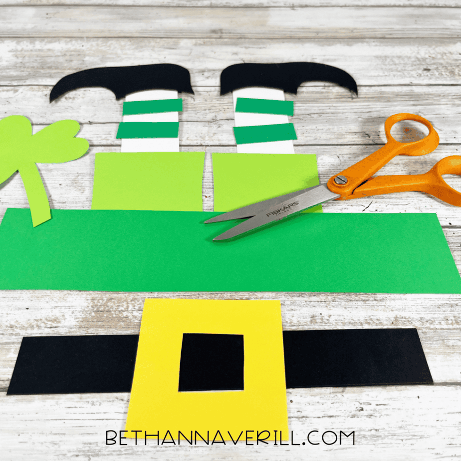 Cut cardstock pieces arranged on a table including striped leg pieces, black shoe shapes, a shamrock shape, and a yellow square frame.