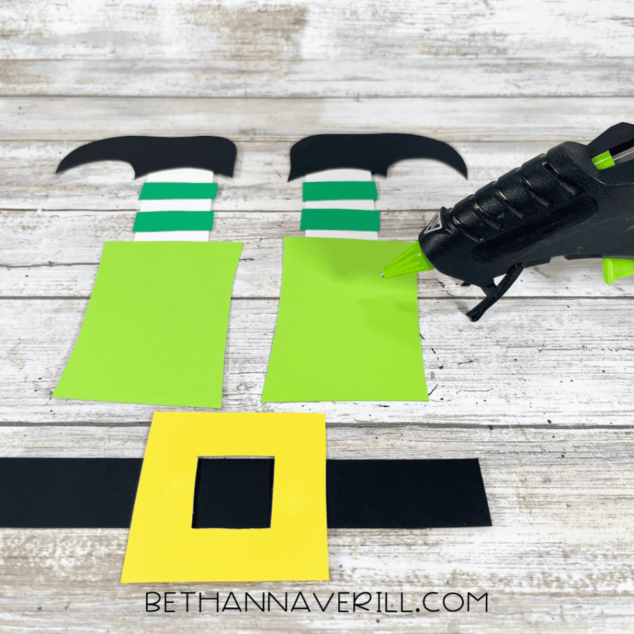 A hot glue gun applying glue to green cardstock rectangles attached to striped leg pieces and black shoe shapes.