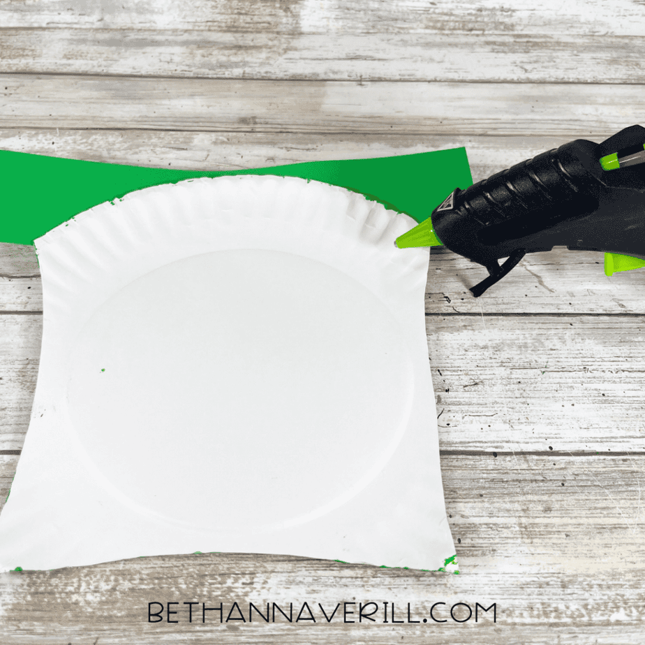 Two assembled striped leg pieces with wooden craft sticks attached being glued to the back of a painted green paper plate piece.