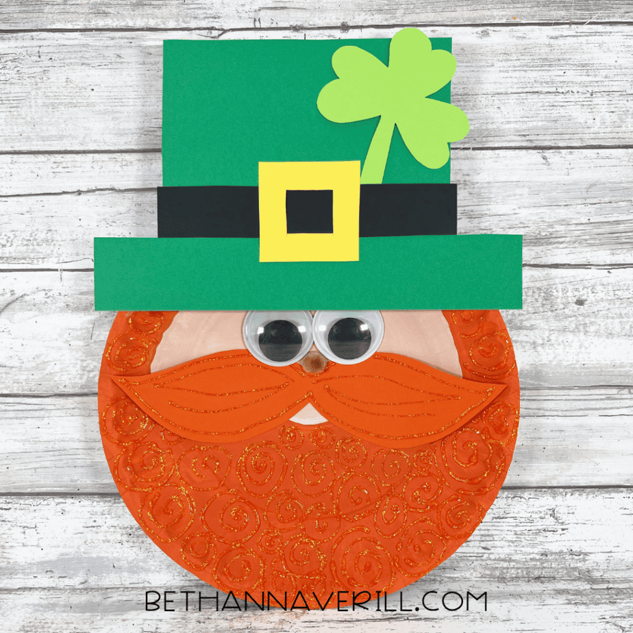 The completed paper plate craft showing a face with googly eyes, an orange mustache, a green hat with a yellow buckle, and orange glitter swirls on the plate.