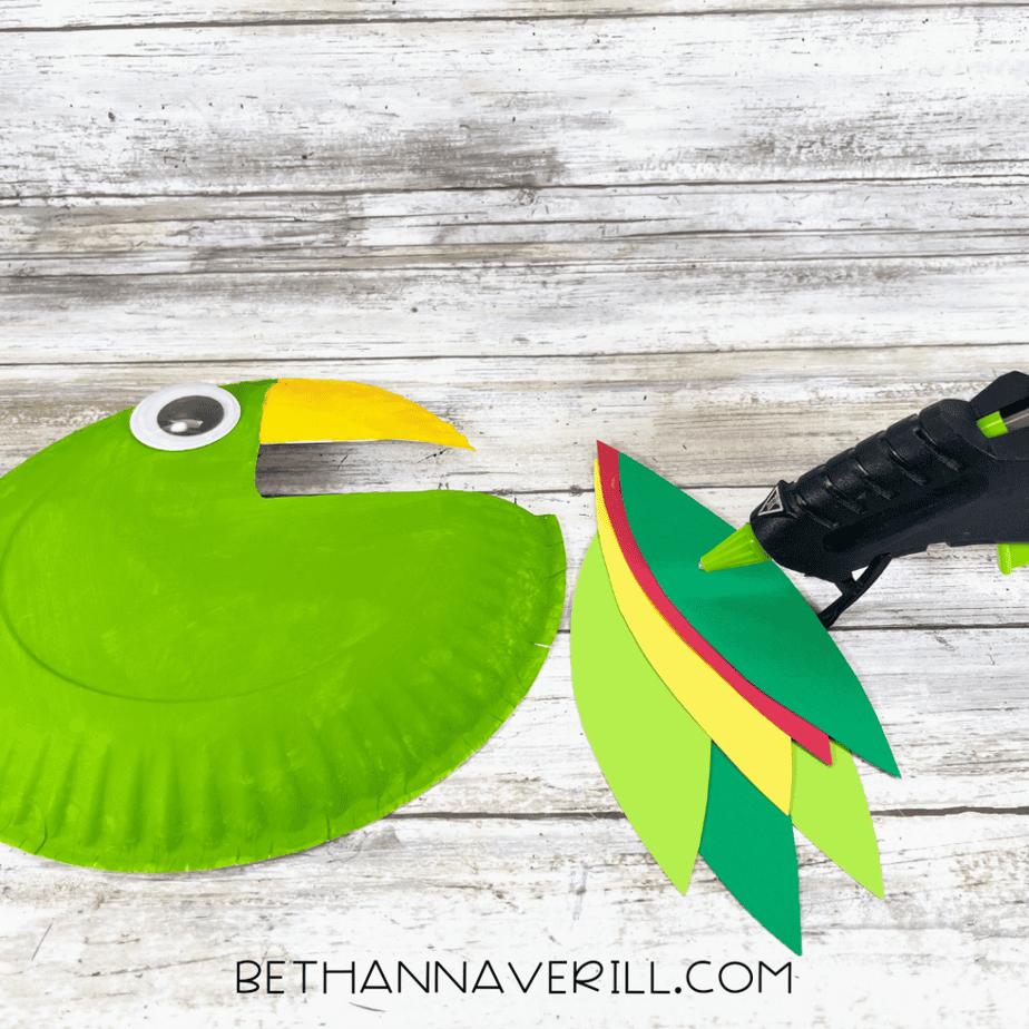Layered cardstock tail feather shapes being glued together next to the green painted paper plate.