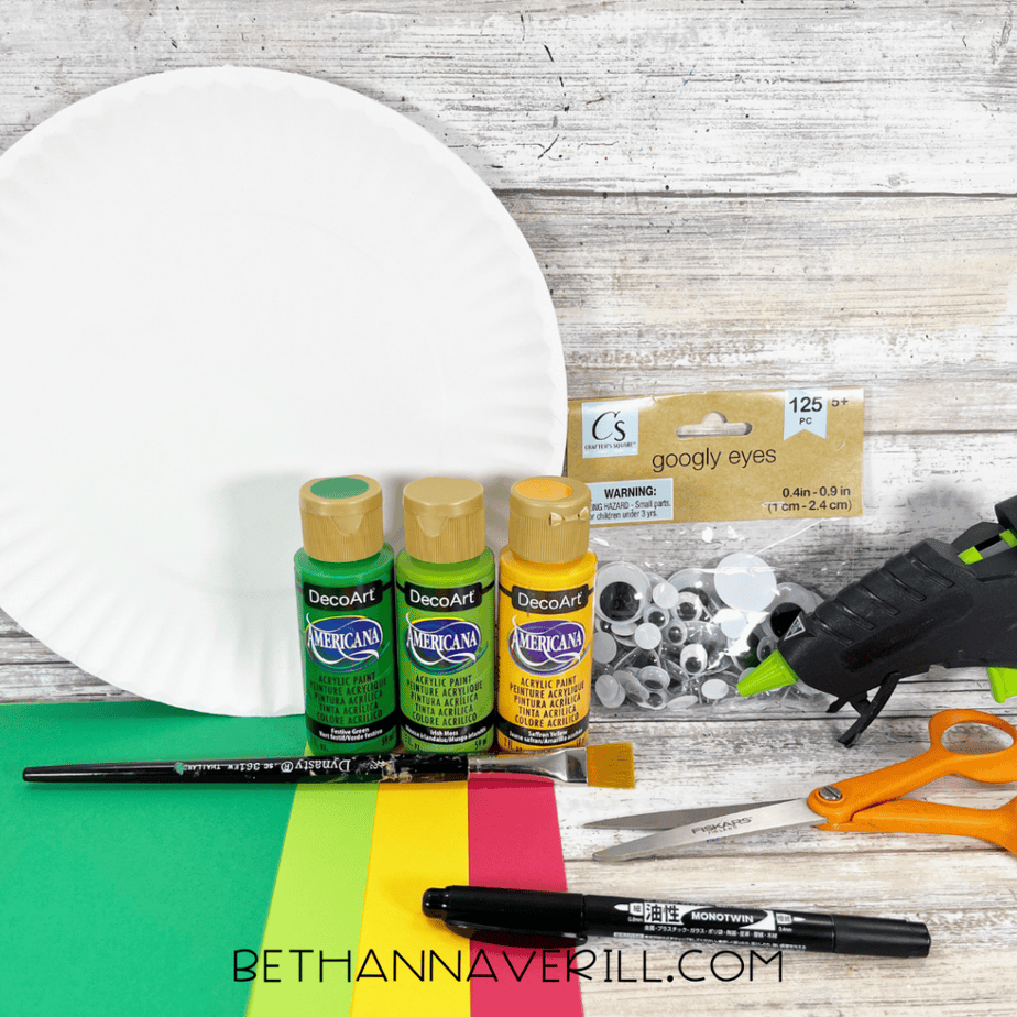 White paper plate, three bottles of acrylic paint (green, light green, yellow), googly eyes package, hot glue gun, scissors, paintbrush, pencil, black marker, and sheets of green, yellow, and red cardstock on a wood surface.
