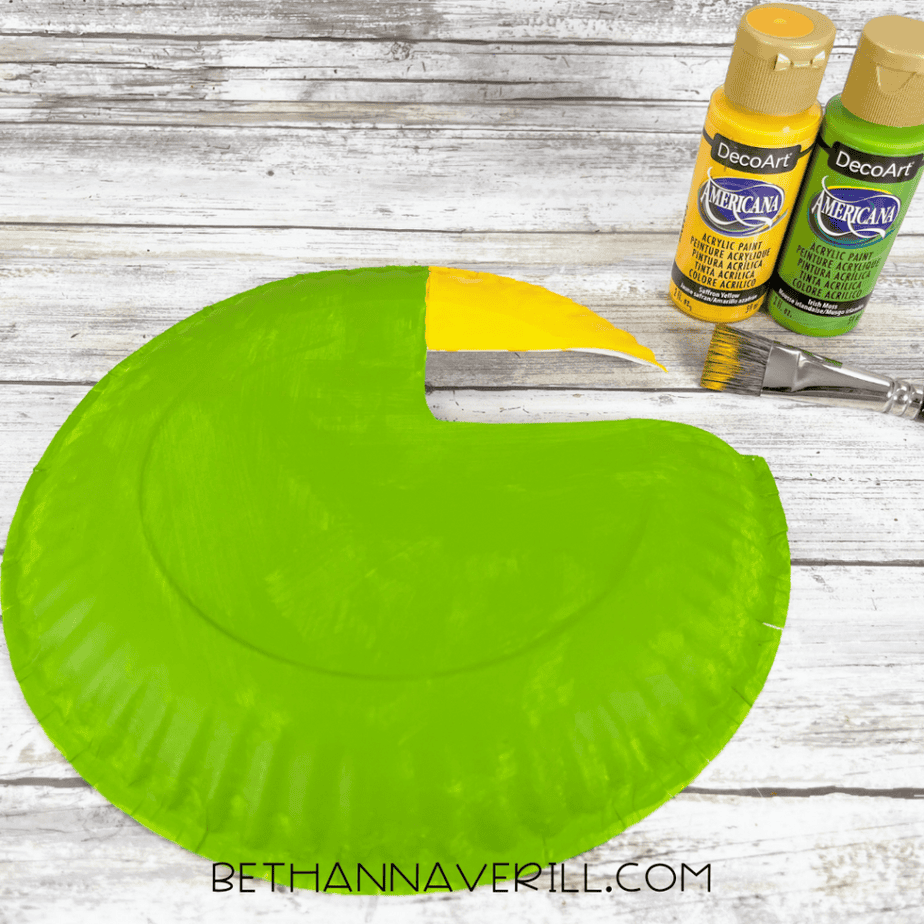 Paper plate painted green with the beak area painted yellow. Two bottles of paint and a paintbrush are next to it.