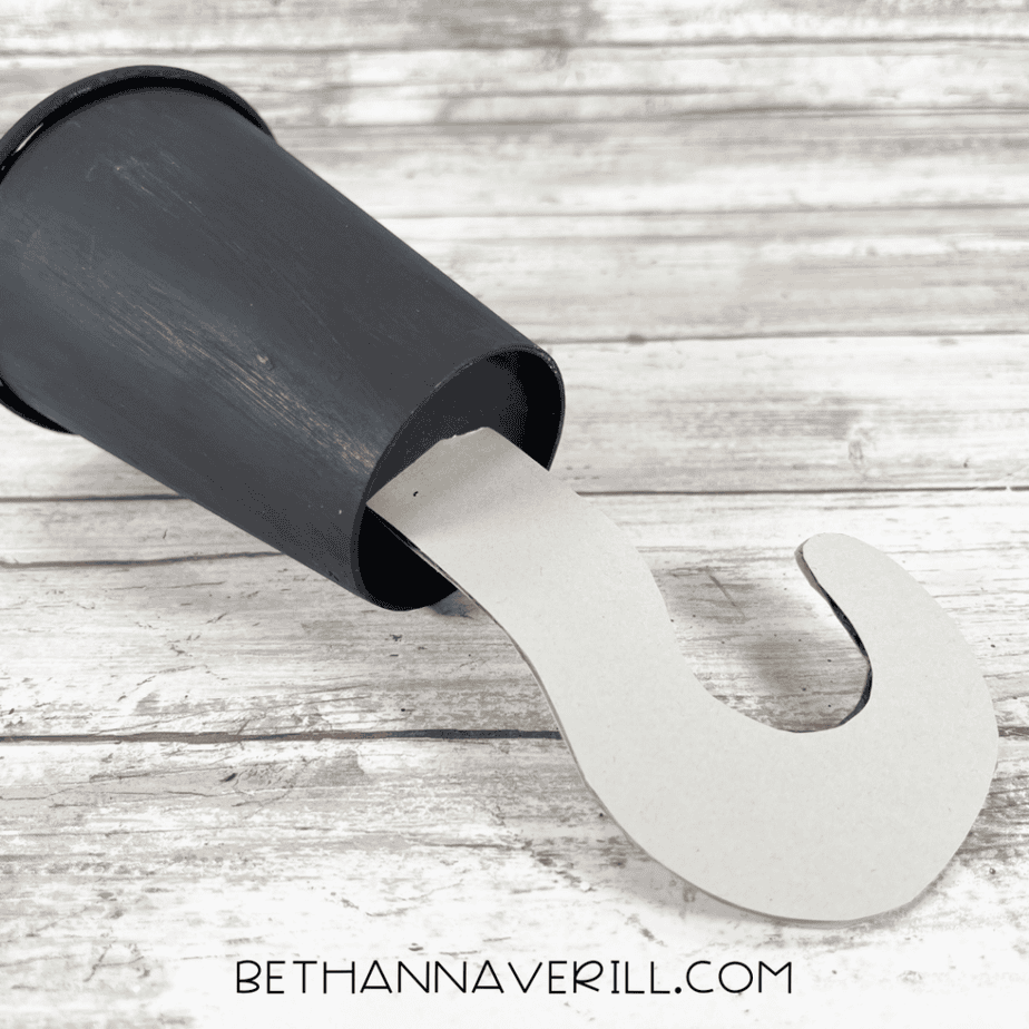 Gray hook-shaped piece inserted into the opening of a black paper cup lying on its side on a wood plank background.