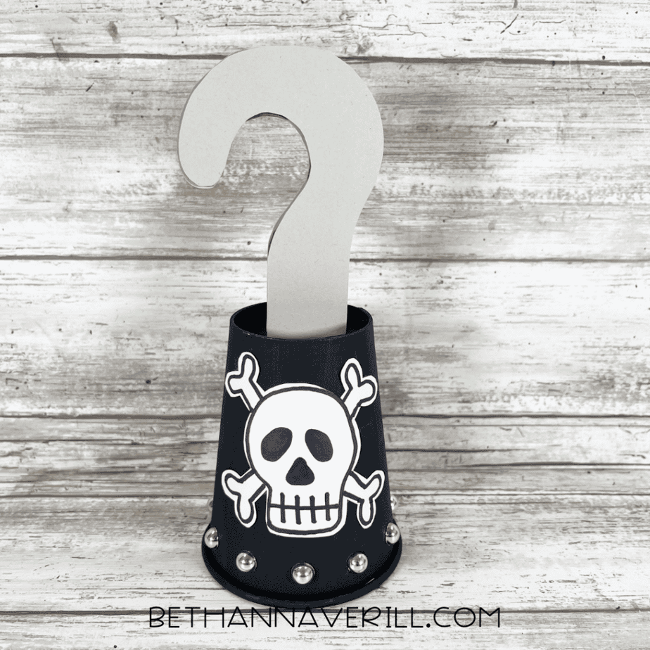 Finished pirate hook craft made from a black paper cup with a gray hook inserted into the top, a white skull and crossbones on the front, and silver rhinestones around the bottom edge.