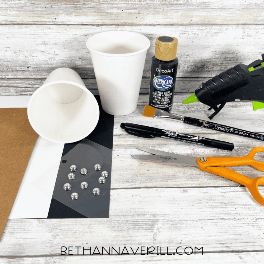 Craft supplies including two white paper cups, black acrylic paint, a paintbrush, scissors, a black marker, a hot glue gun, rhinestones, and pieces of cardstock on a wood background.
