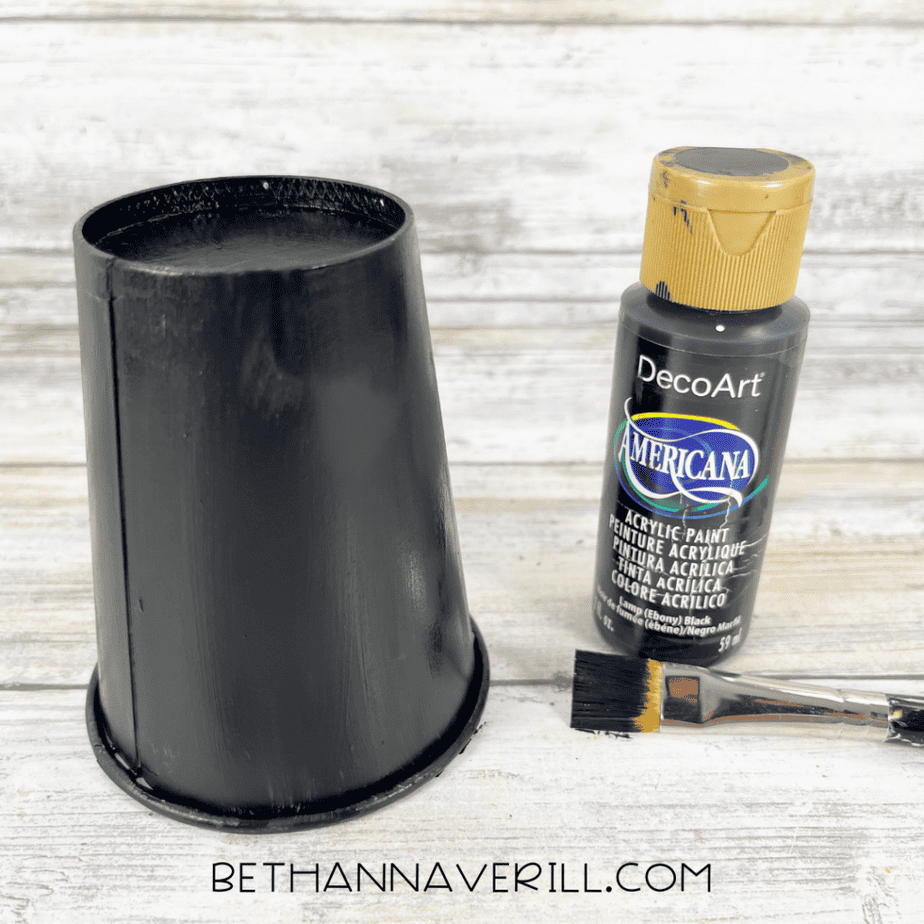 Paper cup painted black next to a bottle of black acrylic paint and a paintbrush.