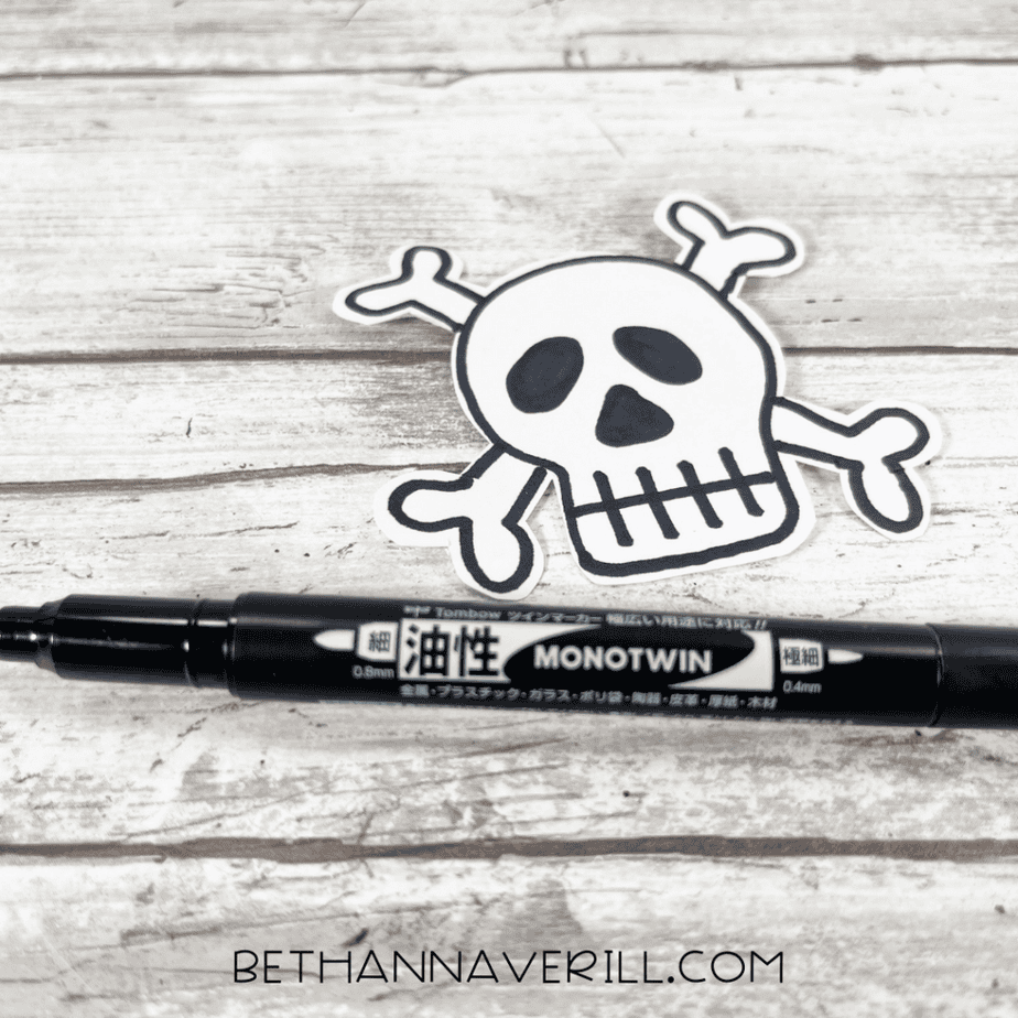Black marker next to a white paper skull and crossbones cutout on a wood plank background.