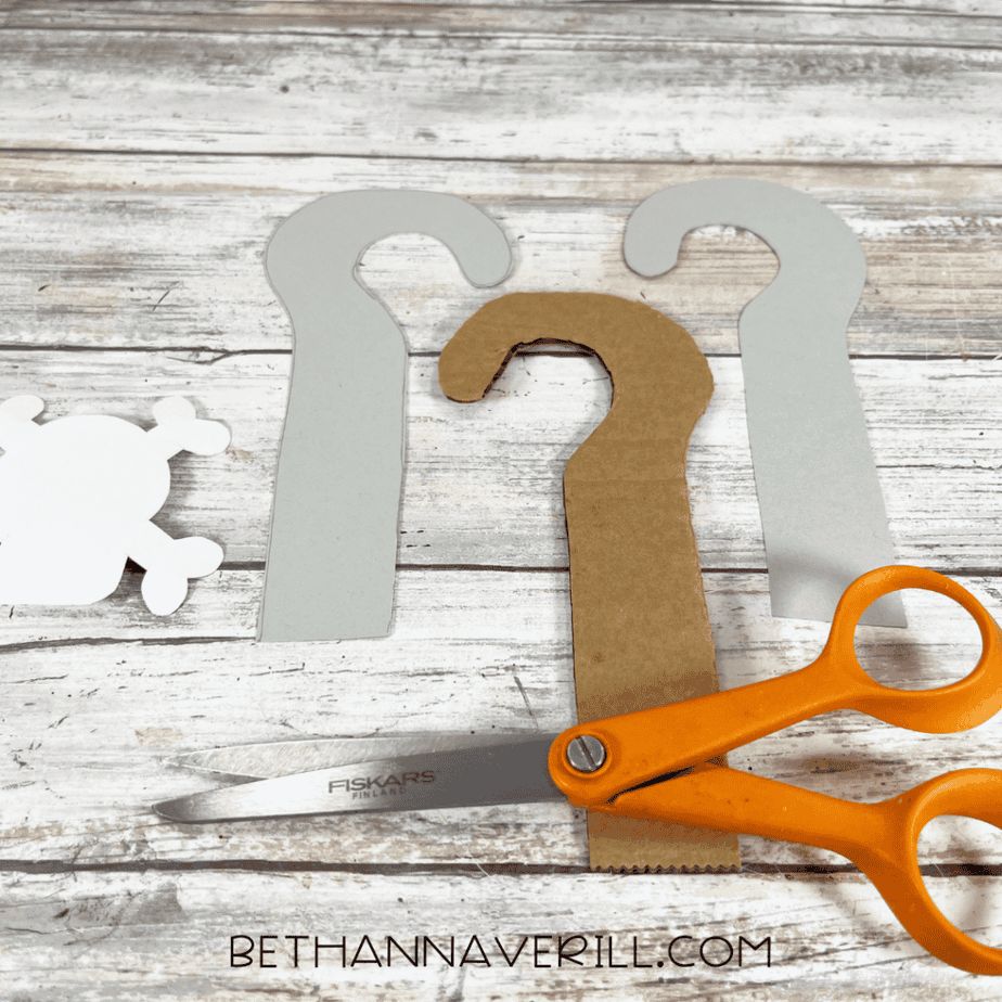 Two gray hook-shaped pieces, one brown cardboard hook-shaped piece, part of a white skull cutout, and orange scissors on a wood plank background.