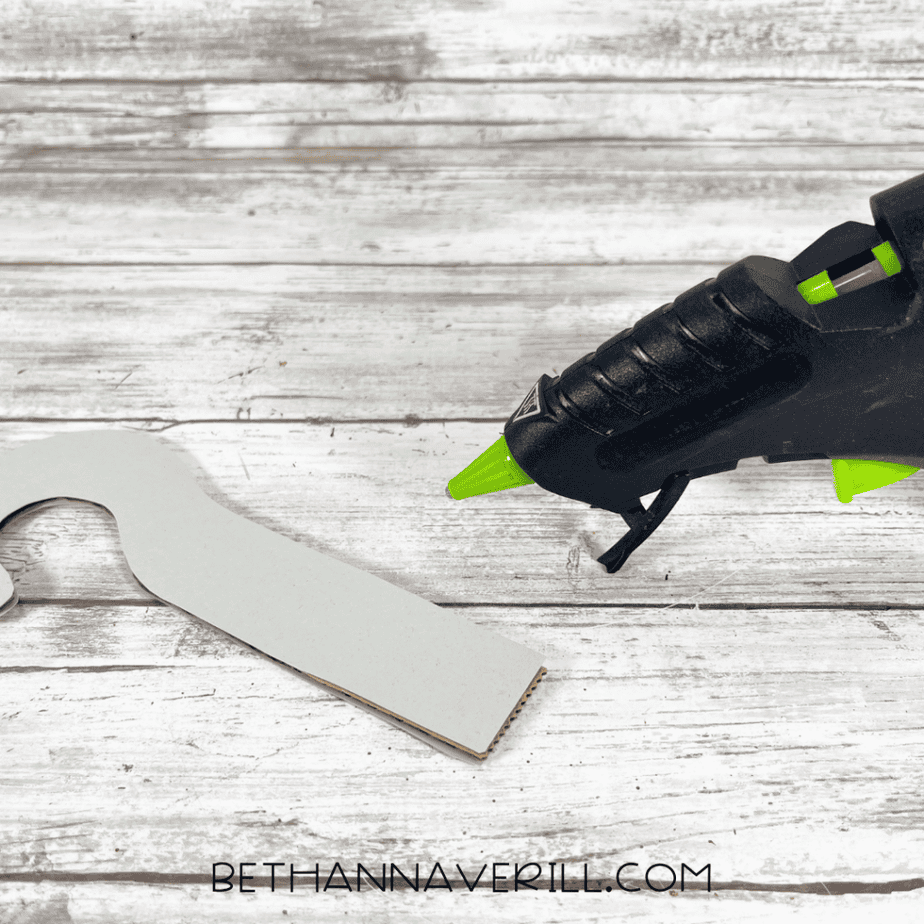 Gray hook-shaped piece with cardboard backing next to a black hot glue gun on a wood plank background.