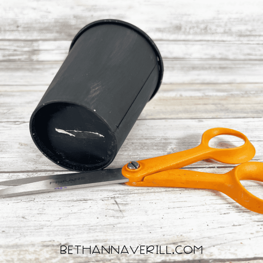 Black paper cup lying on its side next to orange scissors on a wood plank background.
