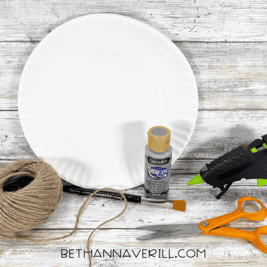 Paper plate, twine, gray acrylic paint bottle, paintbrush, scissors, and hot glue gun on a wooden surface.