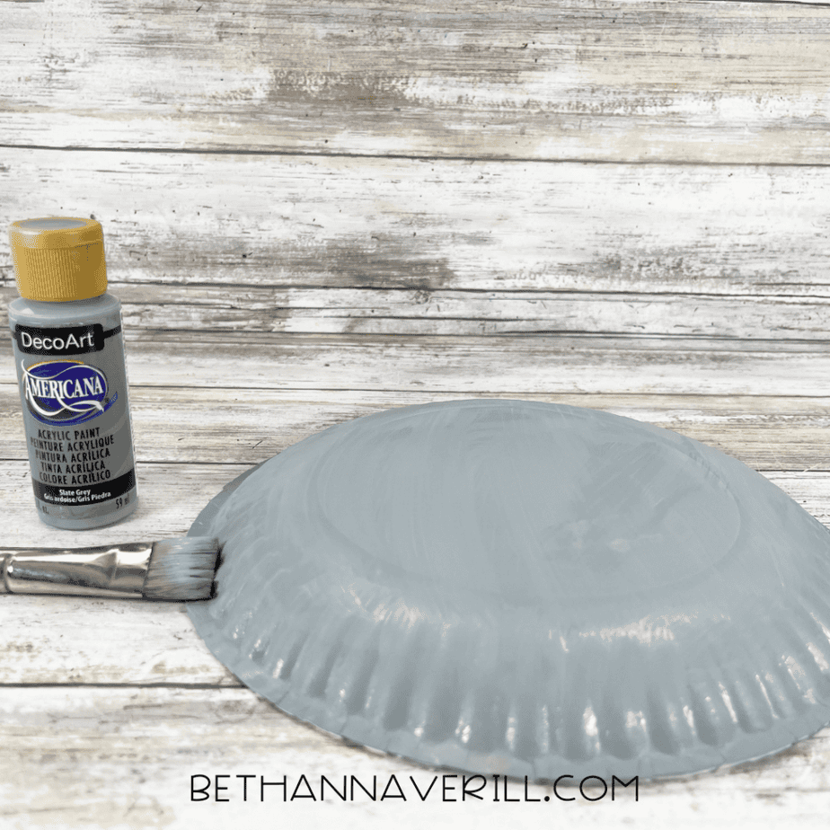 Upside-down paper plate being painted gray with a paintbrush beside a bottle of gray acrylic paint.