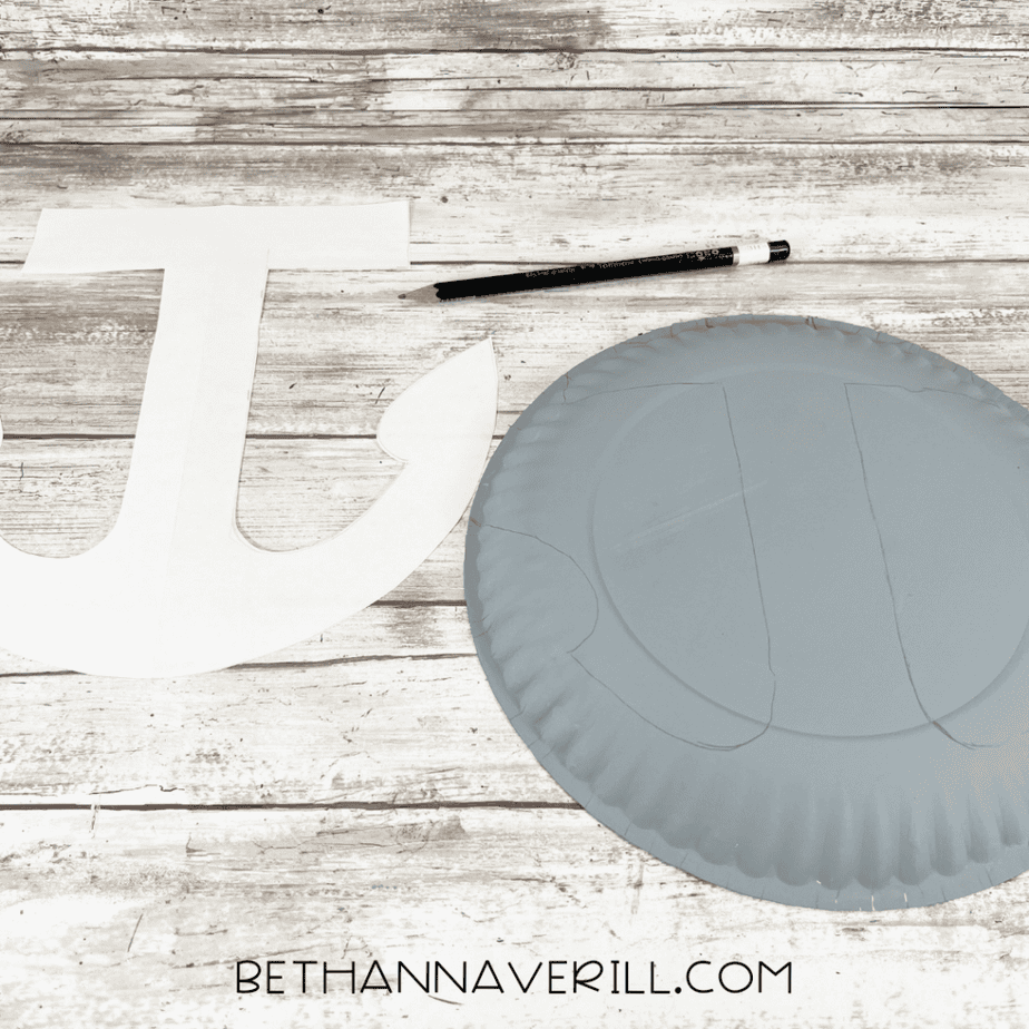 Gray-painted paper plate with anchor shape traced on the surface next to a paper anchor template and pencil.