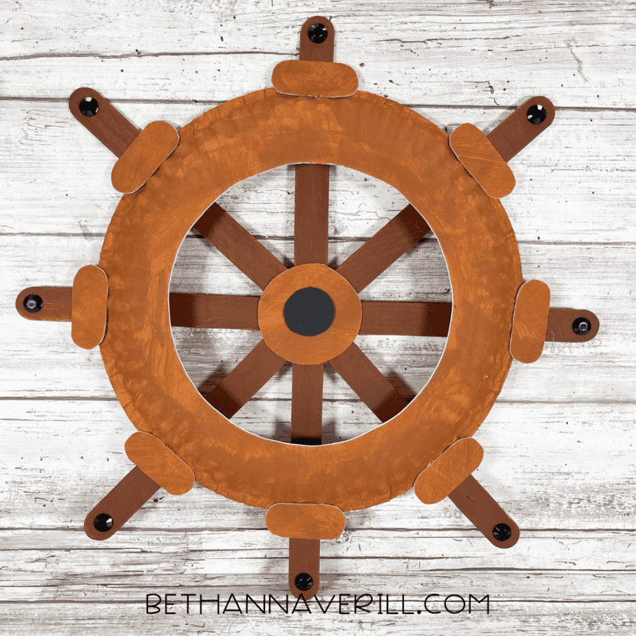Finished paper plate craft shaped like a ship wheel made with painted craft sticks and adhesive gem stickers.
