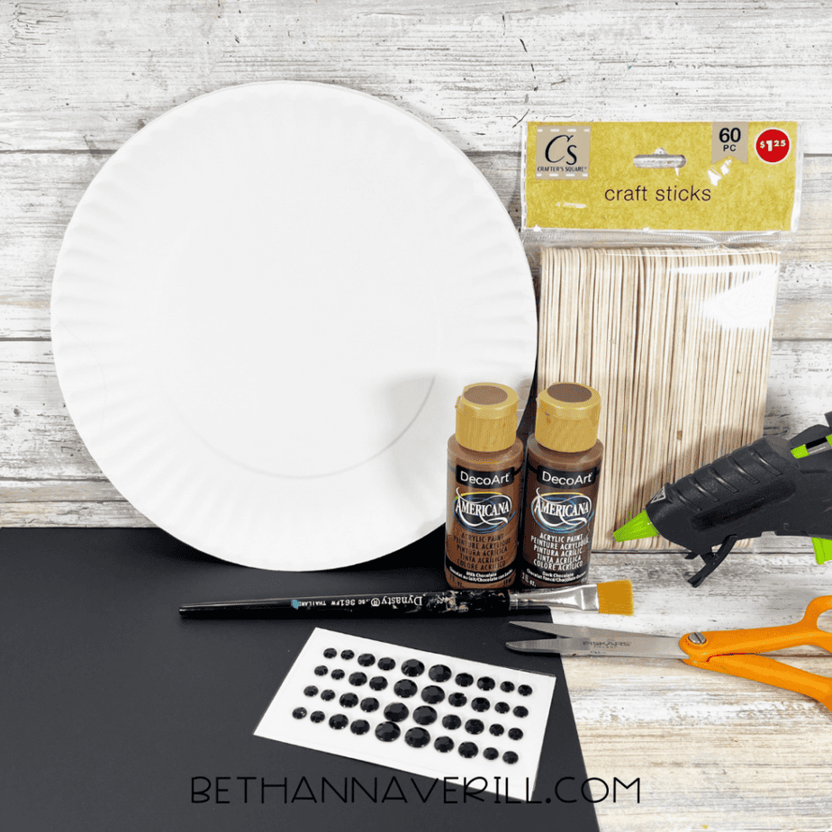 Paper plate, pack of wooden craft sticks, two bottles of brown acrylic paint, paintbrush, scissors, glue gun, and adhesive gem stickers on a wooden background.