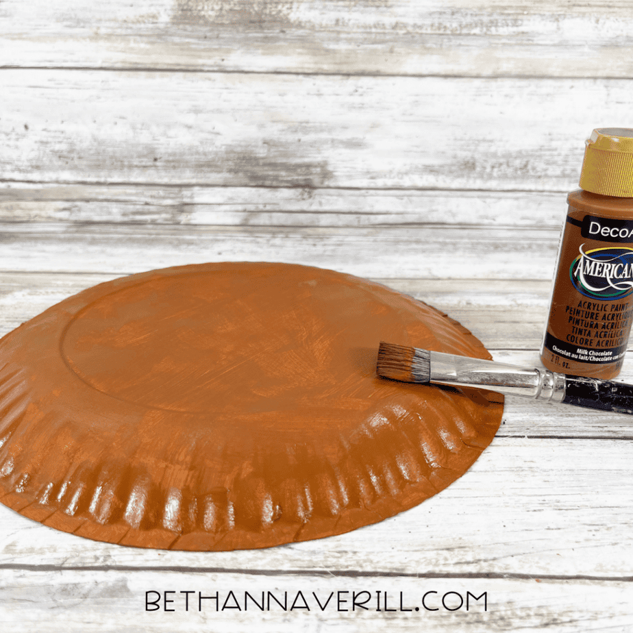 Upside-down paper plate partially painted brown with a paintbrush and bottle of brown acrylic paint beside it.