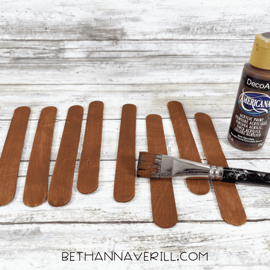 Several wooden craft sticks laid out in a row with brown paint being applied using a paintbrush.