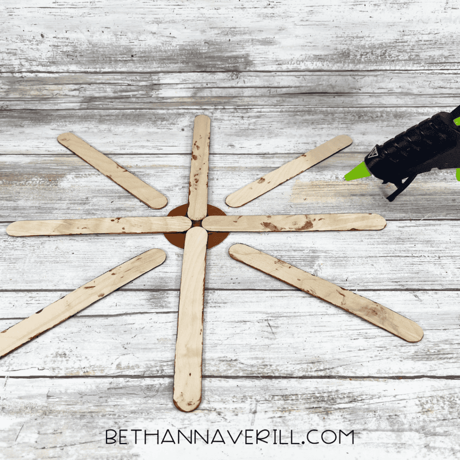 Wooden craft sticks arranged in a circular pattern around a small round cardboard piece with a glue gun nearby.