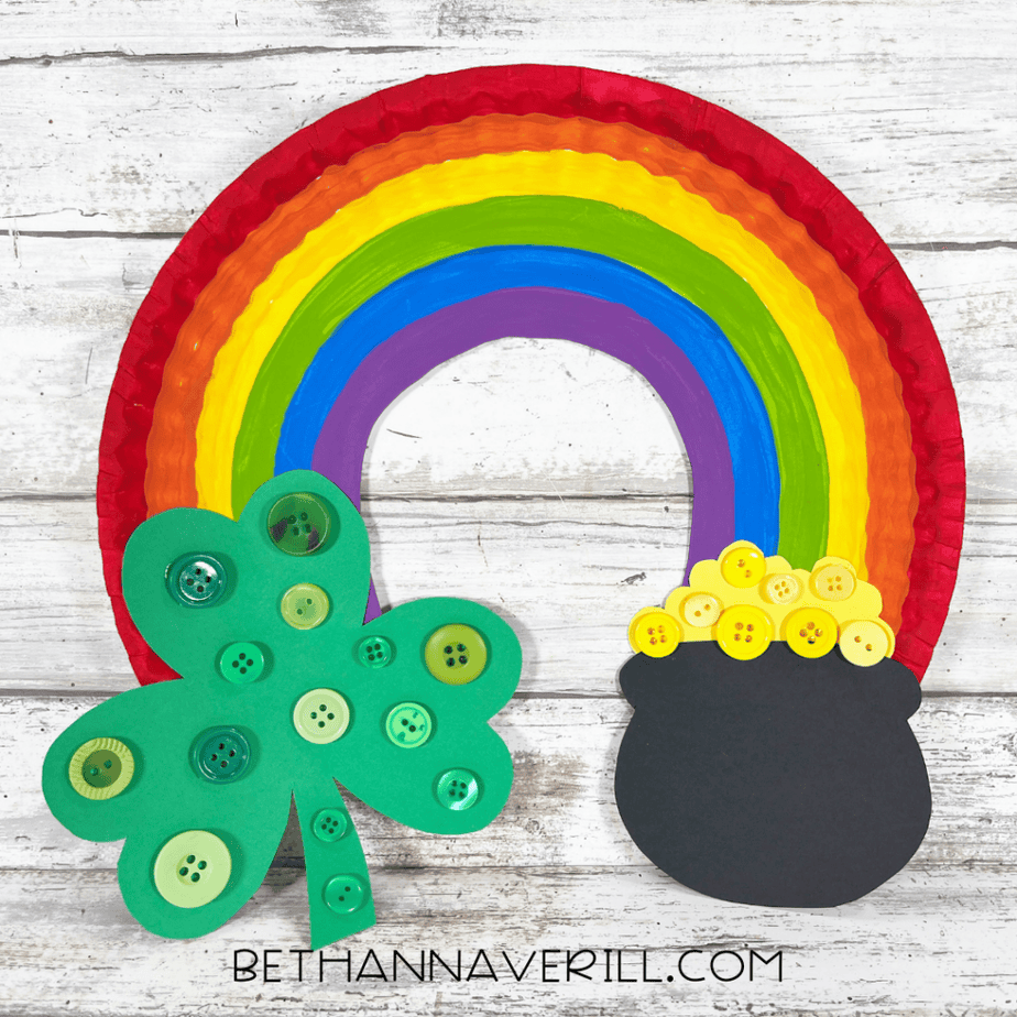 A completed paper plate rainbow craft displayed with a green button-decorated shamrock on the left and a black pot with yellow buttons on the right.