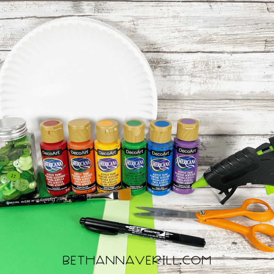 Craft supplies arranged on a wooden surface including a white paper plate, six bottles of acrylic paint in rainbow colors, green buttons in a jar, green and light green cardstock, a paintbrush, black marker, scissors, and a hot glue gun.