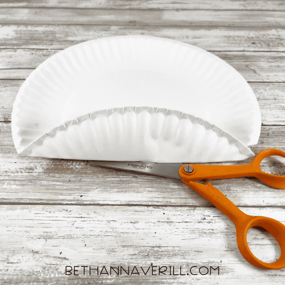 A white paper plate folded in half resting on a wooden surface with orange-handled scissors placed underneath.