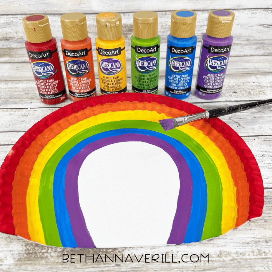A half paper plate painted in rainbow stripes with red, orange, yellow, green, blue, and purple bands. Paint bottles and a paintbrush are displayed behind it.