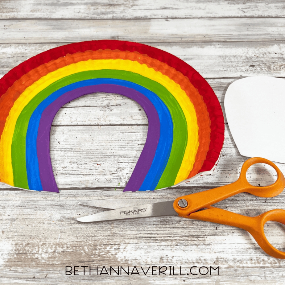 The center of the painted rainbow paper plate has been cut out, leaving a rainbow arch shape. Orange-handled scissors are placed beside it.