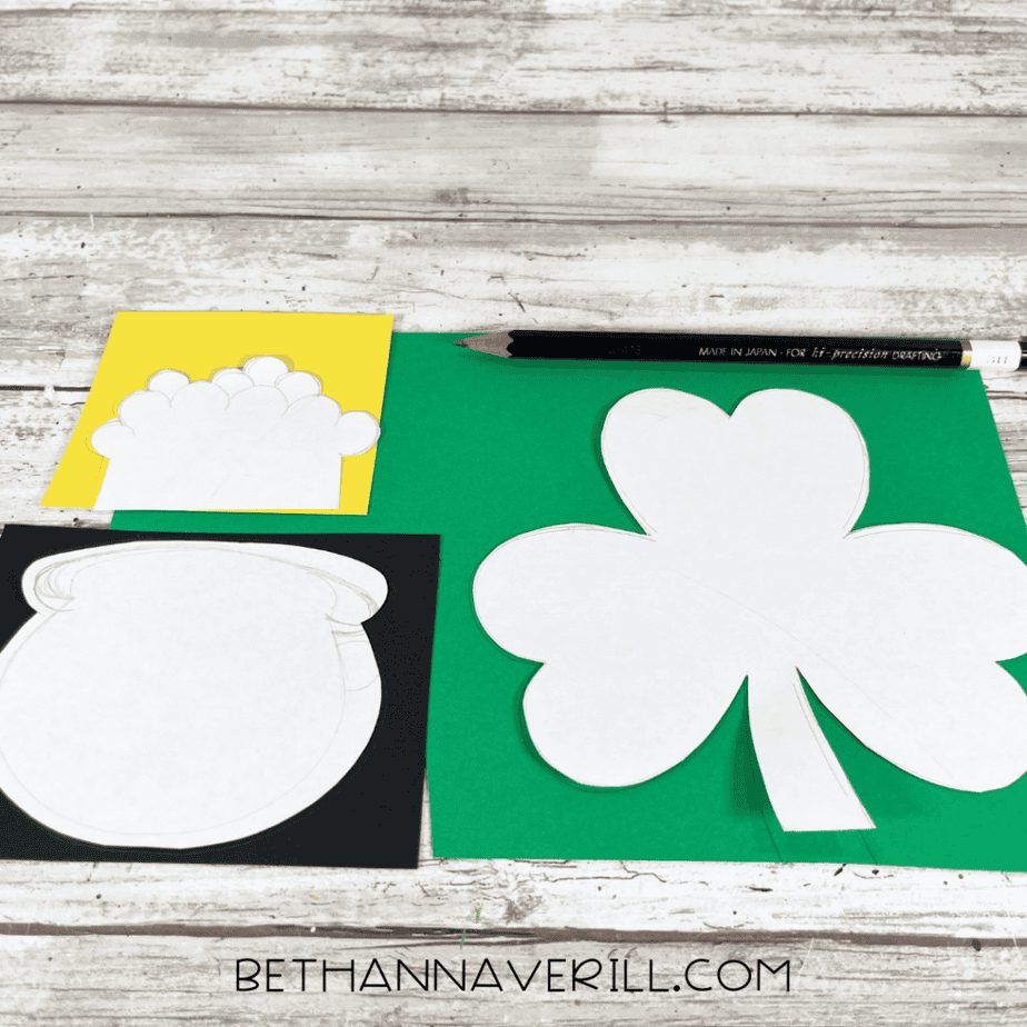 Paper pattern templates for a shamrock, pot, and cloud placed on green, black, and yellow cardstock with a pencil on a wooden surface.